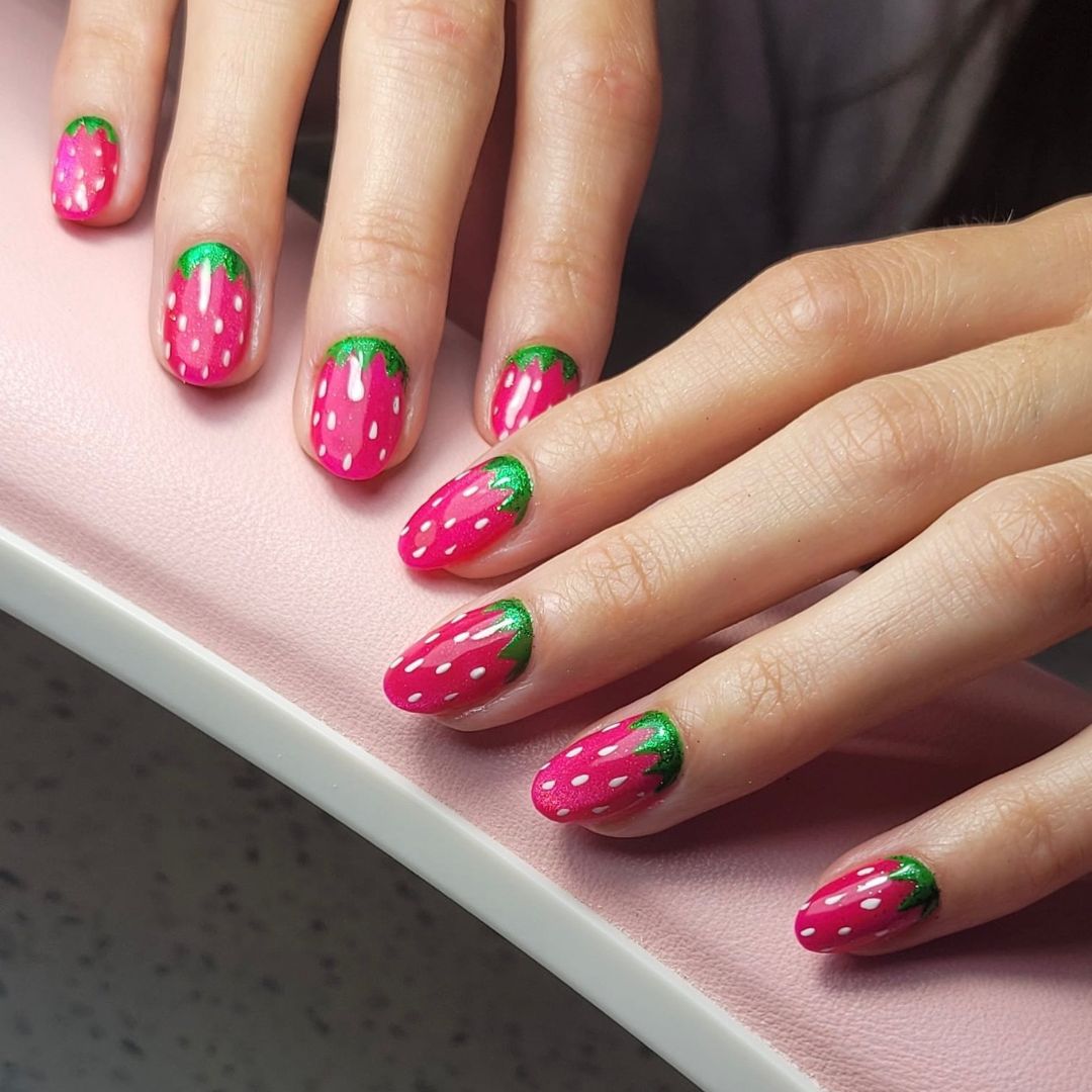 Strawberry-inspired nail art design with bright pink gel polish, white seed detailing, and green leaf accents near the cuticle.